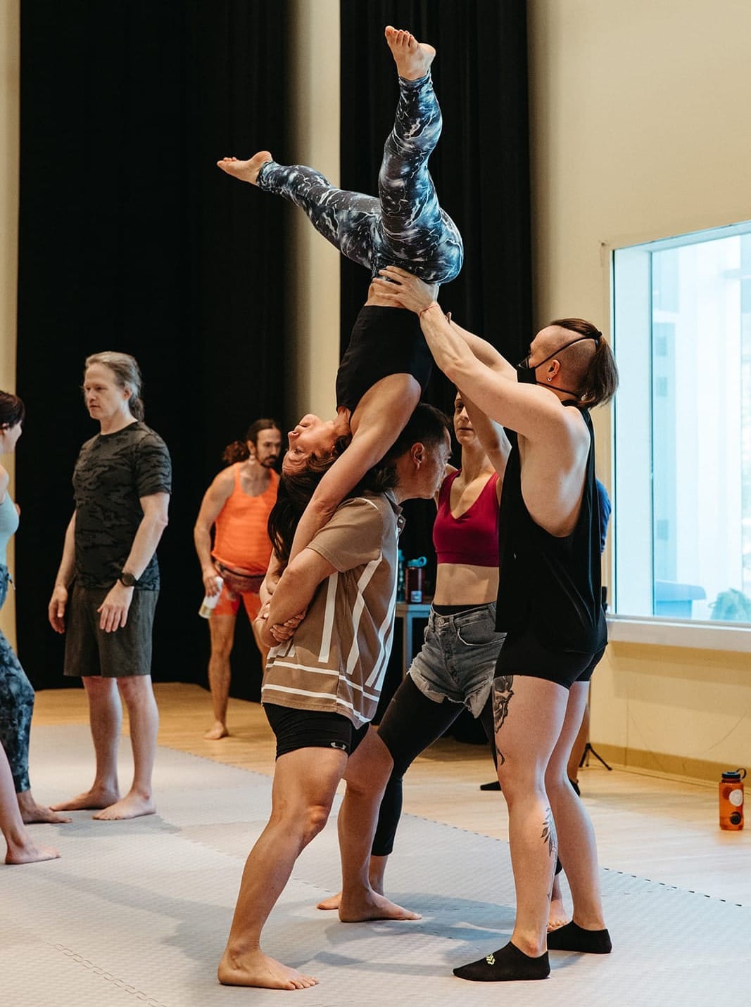 A flyer inverted, balanced on their hands against a base's back, with two spotters standing behind to prevent falls or injuries