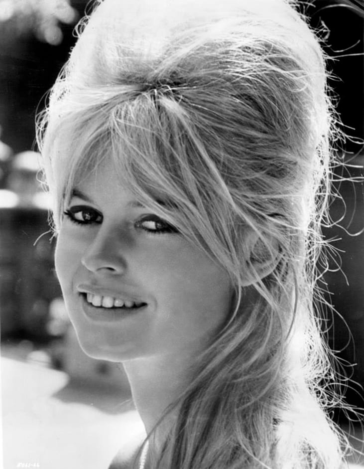 Black and white photo of Brigitte Bardot staring straight into the camera with a slightly-open-mouth smile and wisps of hair in her face, with iconic 1960s black eyeliner