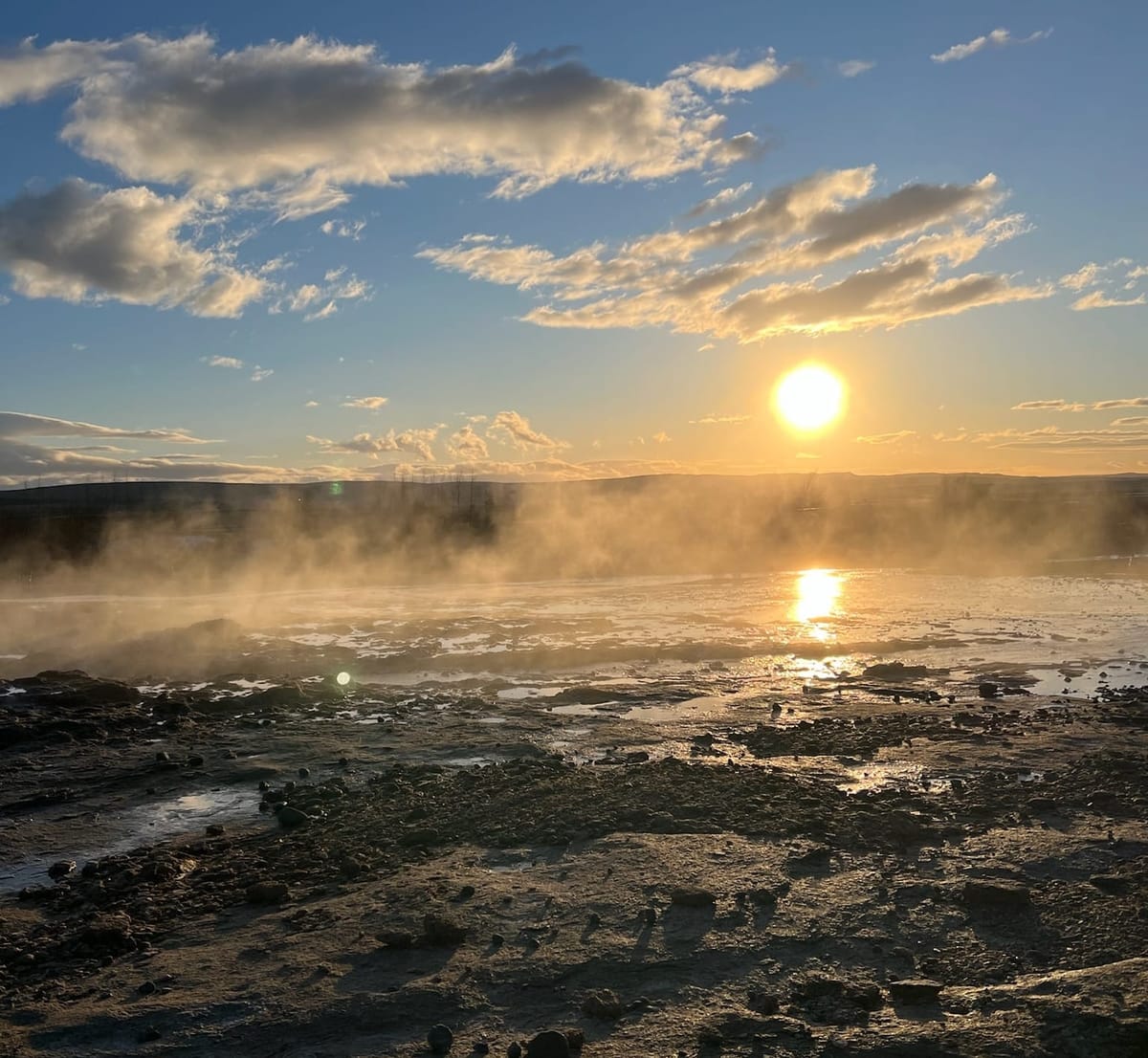 The sun shining over steam from a geysir surrounded by rocks and earth in Iceland.